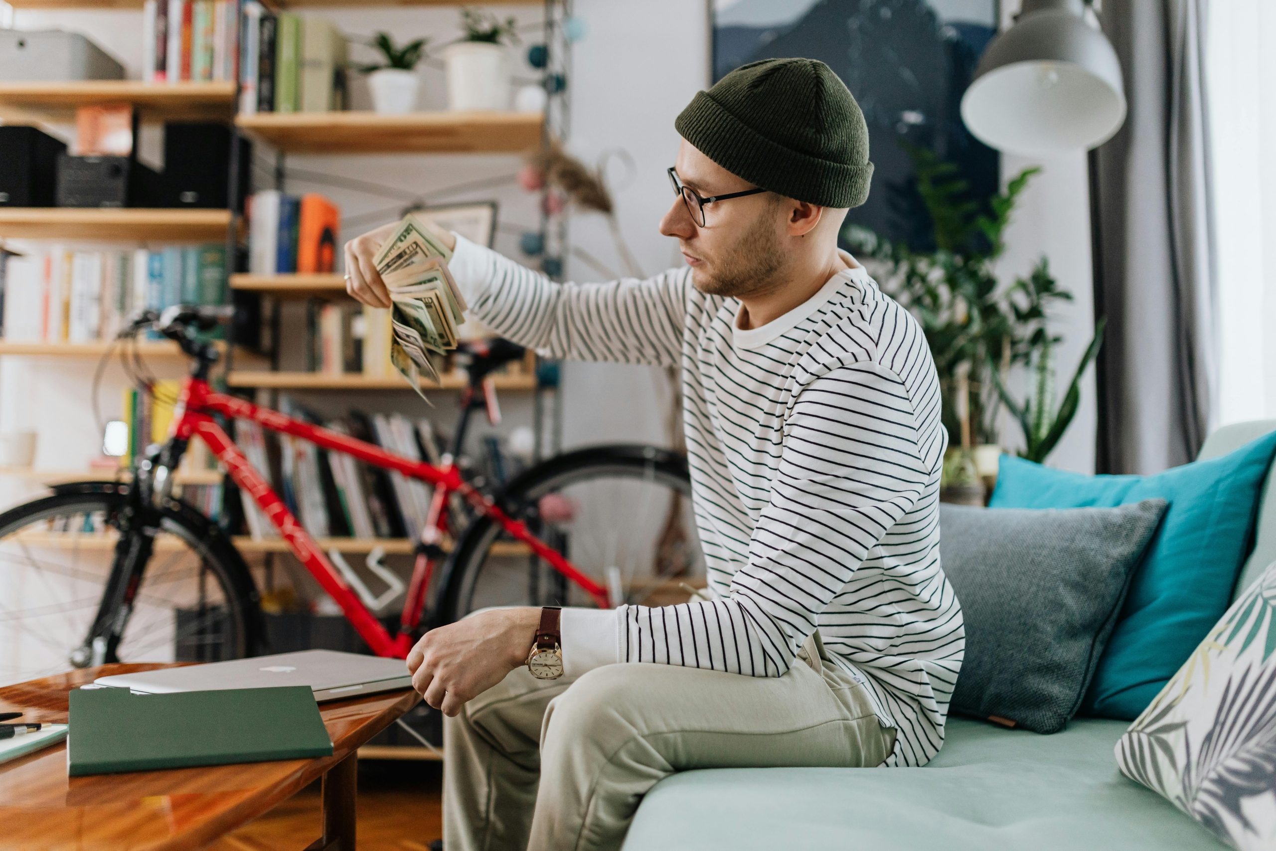Un homme en tenue décontractée comptant de l'argent assis sur un canapé dans un cadre domestique avec un vélo en arrière-plan.