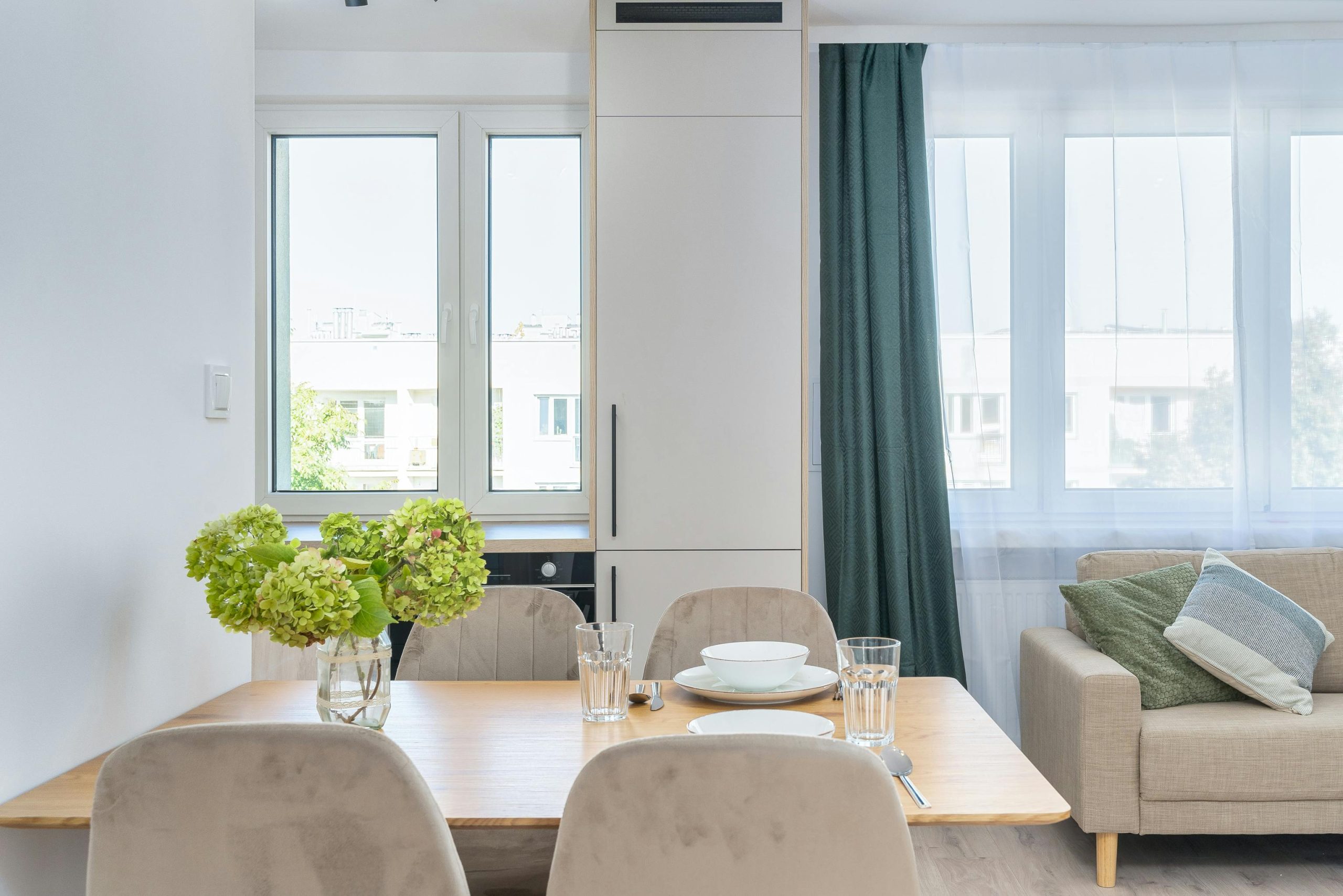 Bright modern dining area with wooden table, hydrangeas, and large windows providing natural light. Salle à manger moderne et lumineuse avec table en bois, hortensias et grandes fenêtres offrant une lumière naturelle.