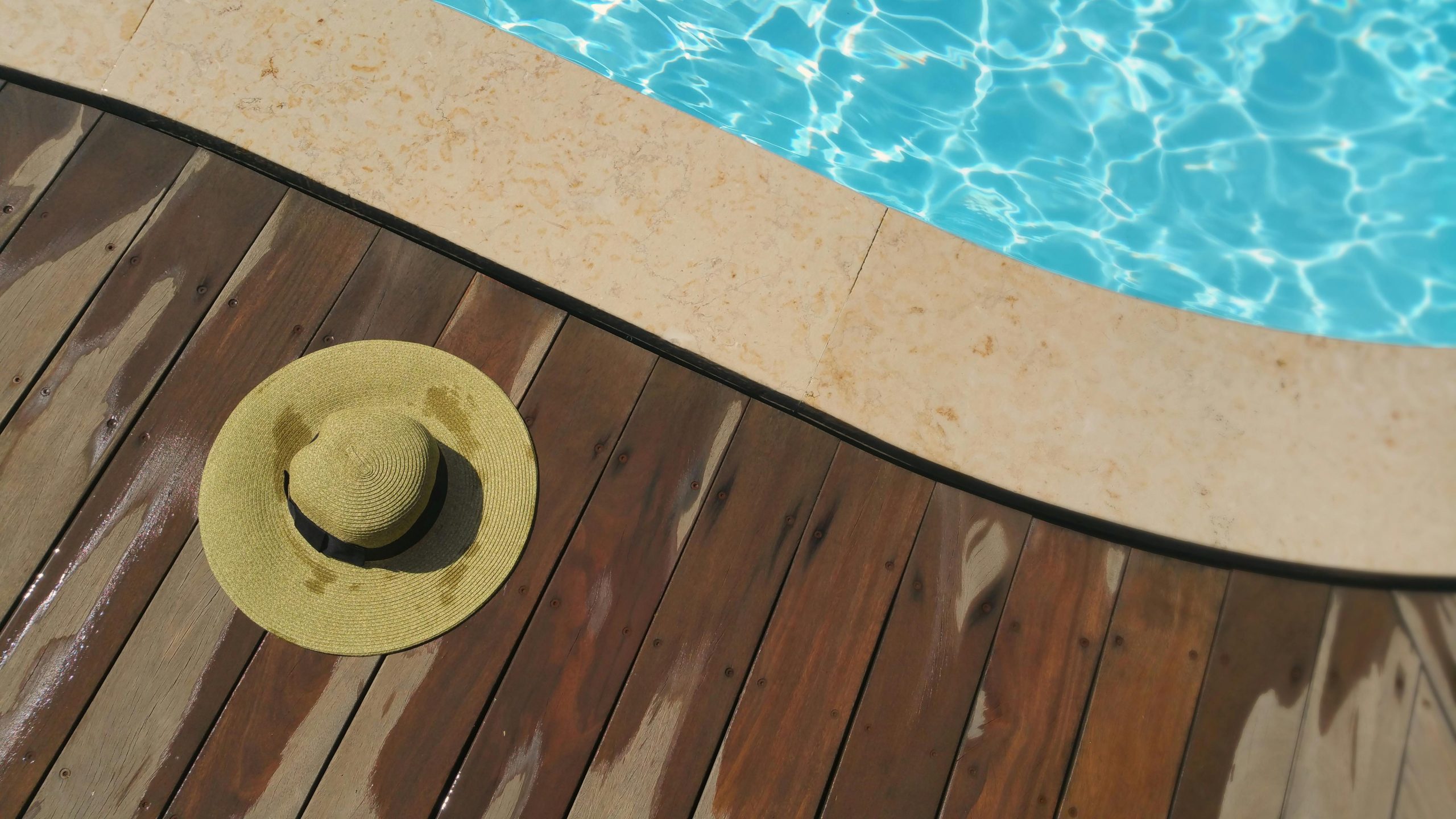 A straw hat rests on a wooden deck beside a sparkling blue swimming pool, perfect for summer relaxation. Un chapeau de paille repose sur une terrasse en bois à côté d'une piscine bleue étincelante, parfait pour la détente estivale.
