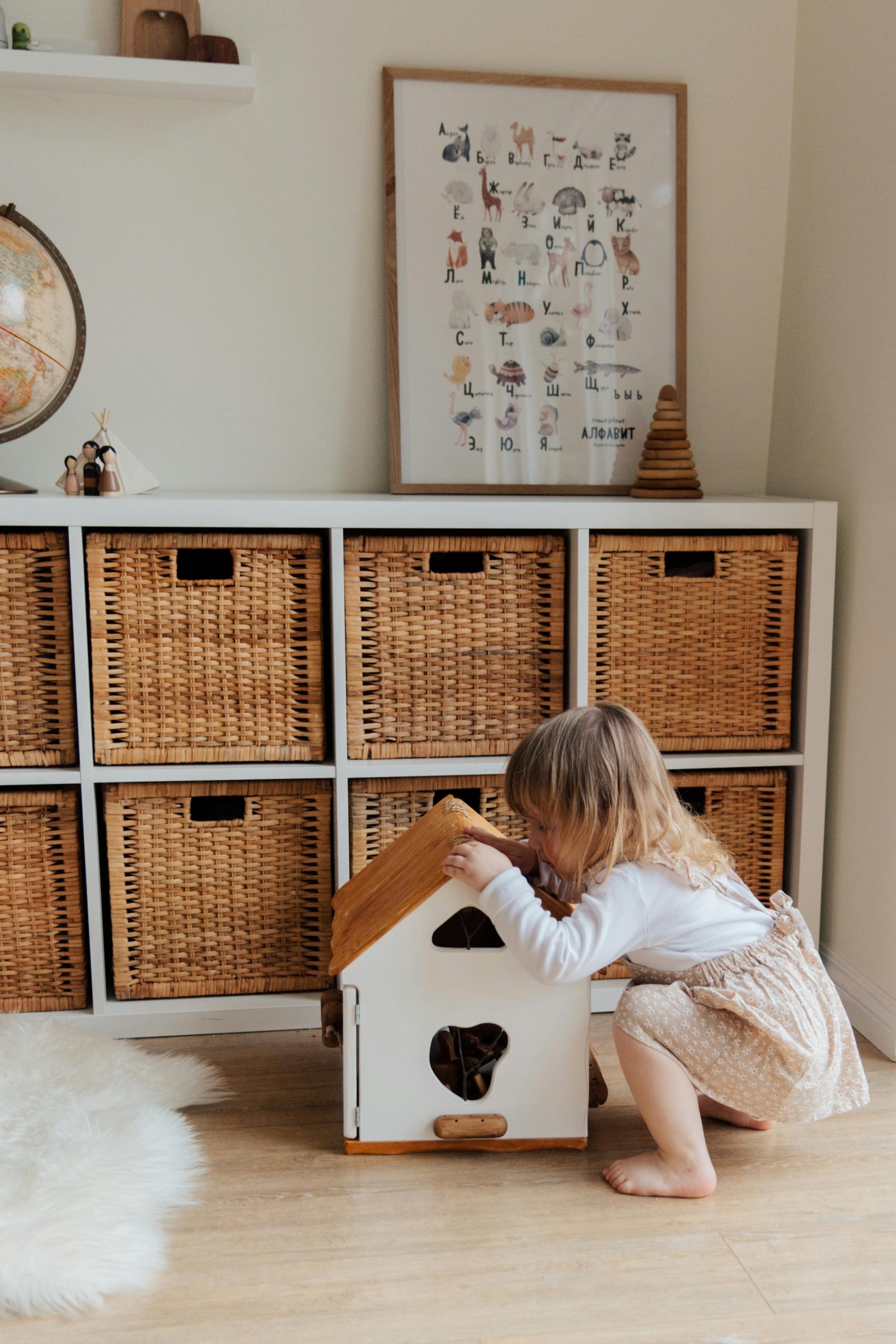 A cute toddler plays with a dollhouse in a cozy room with wooden storage baskets. Un adorable tout-petit joue avec une maison de poupée dans une pièce confortable avec des paniers de rangement en bois.