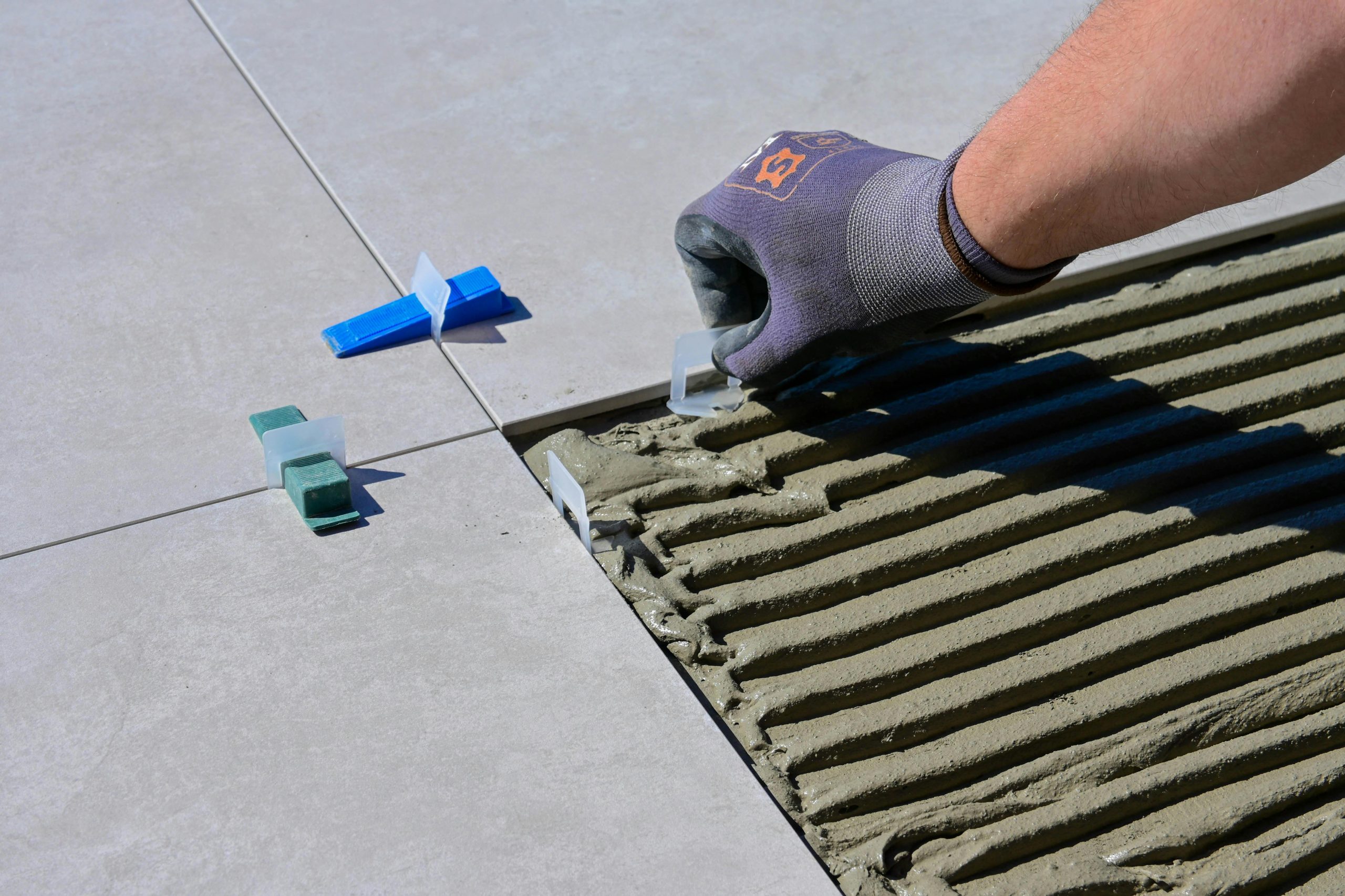 Detailed view of gloved hand aligning tiles with levelers and spacers during construction. Vue détaillée d'une main gantée alignant les carreaux avec des niveaux et des espaceurs lors de la construction.