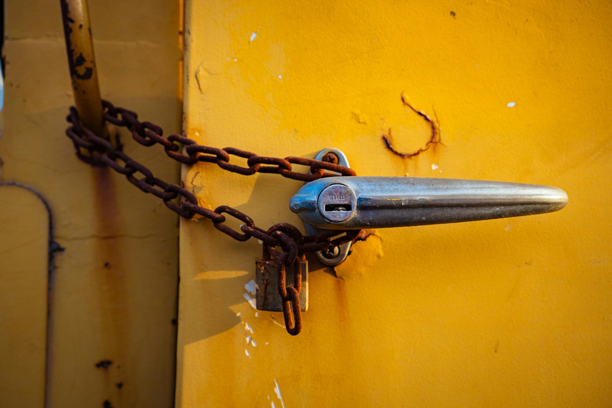 Close-up of a rusty chain securing a chrome handle on an old yellow metal door. Gros plan d'une chaîne rouillée sécurisant une poignée chromée sur une vieille porte métallique jaune.