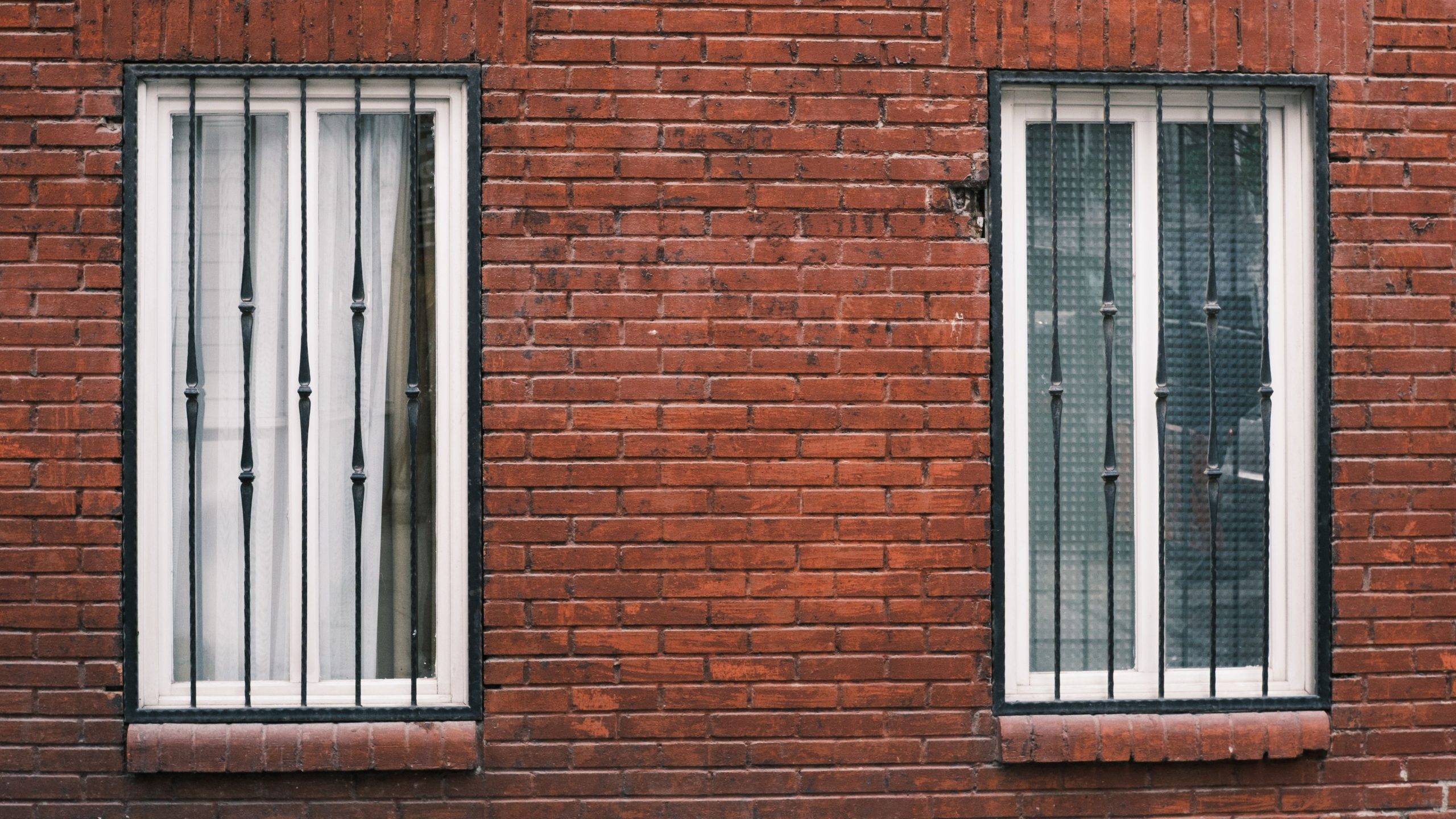 Red brick wall with two barred windows in urban setting. Mur de briques rouges avec deux fenêtres barreaudées en milieu urbain.