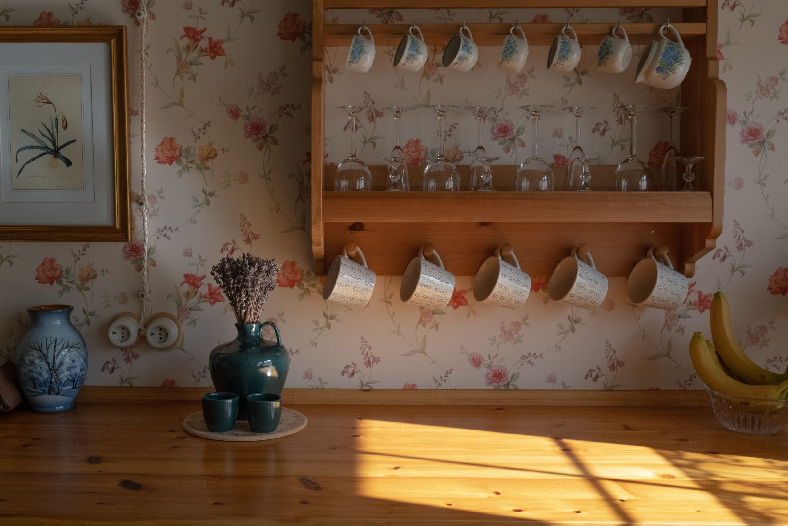 Scène de cuisine vintage avec des tasses florales sur une étagère en bois, la lumière du soleil projetant des ombres chaudes.
